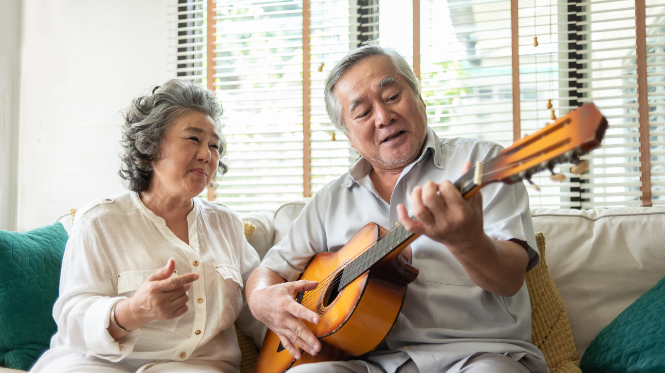 A woman sits next to her husband on the couch while he plays guitar